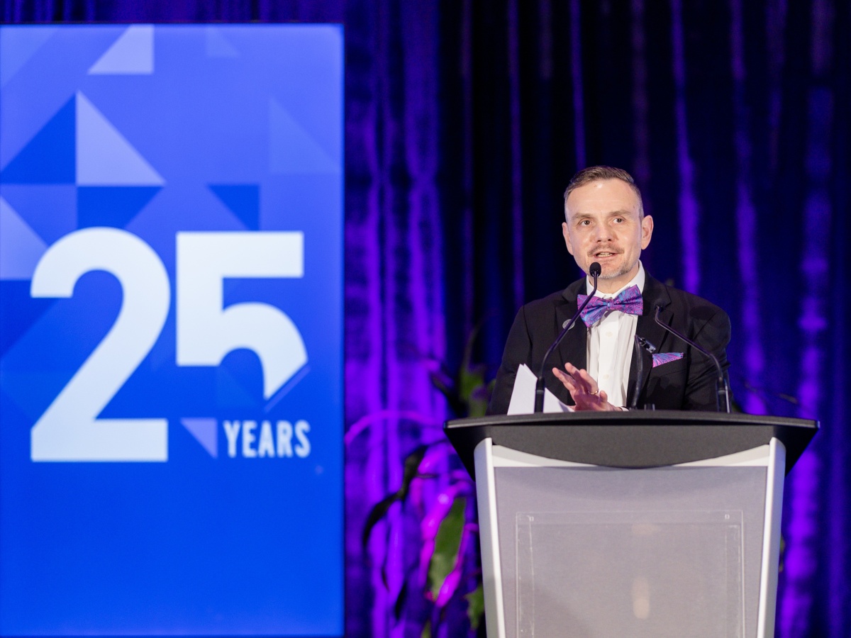 Dr. Geoff Payne in formal wear, standing in front of a podium on a stage. Behind him is a TV screen with a graphic that celebrates 25 years of Michael Smith Health Research BC.