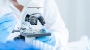 A gloved researcher in a white lab coat uses a pipette to place or collect a sample on a microscope stage in a clean, well‑lit laboratory. The photo is shot closeup, so that the focus is on the researcher's hands and equipment.