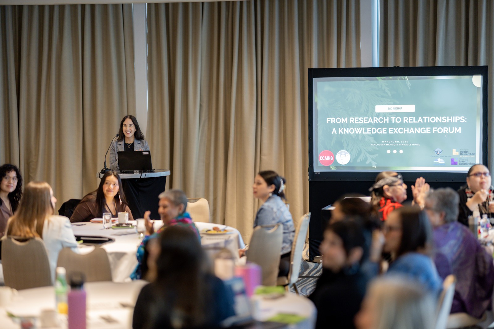 Dr. Krista Stelkia stands at a podium in the room and speaks to a room of seated people at the Knowledge Exchange Forum: From Research to Relationships, hosted by BC NEIHR. On the right, a slideshow presentation with the event name is visible.