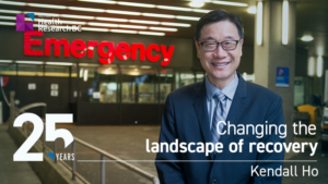 Feature image - Dr. Kendall Ho i standing in front of the outside of an Emergency department. There's a text overlay that reads: "25 years: Changing the landscape of recovery."
